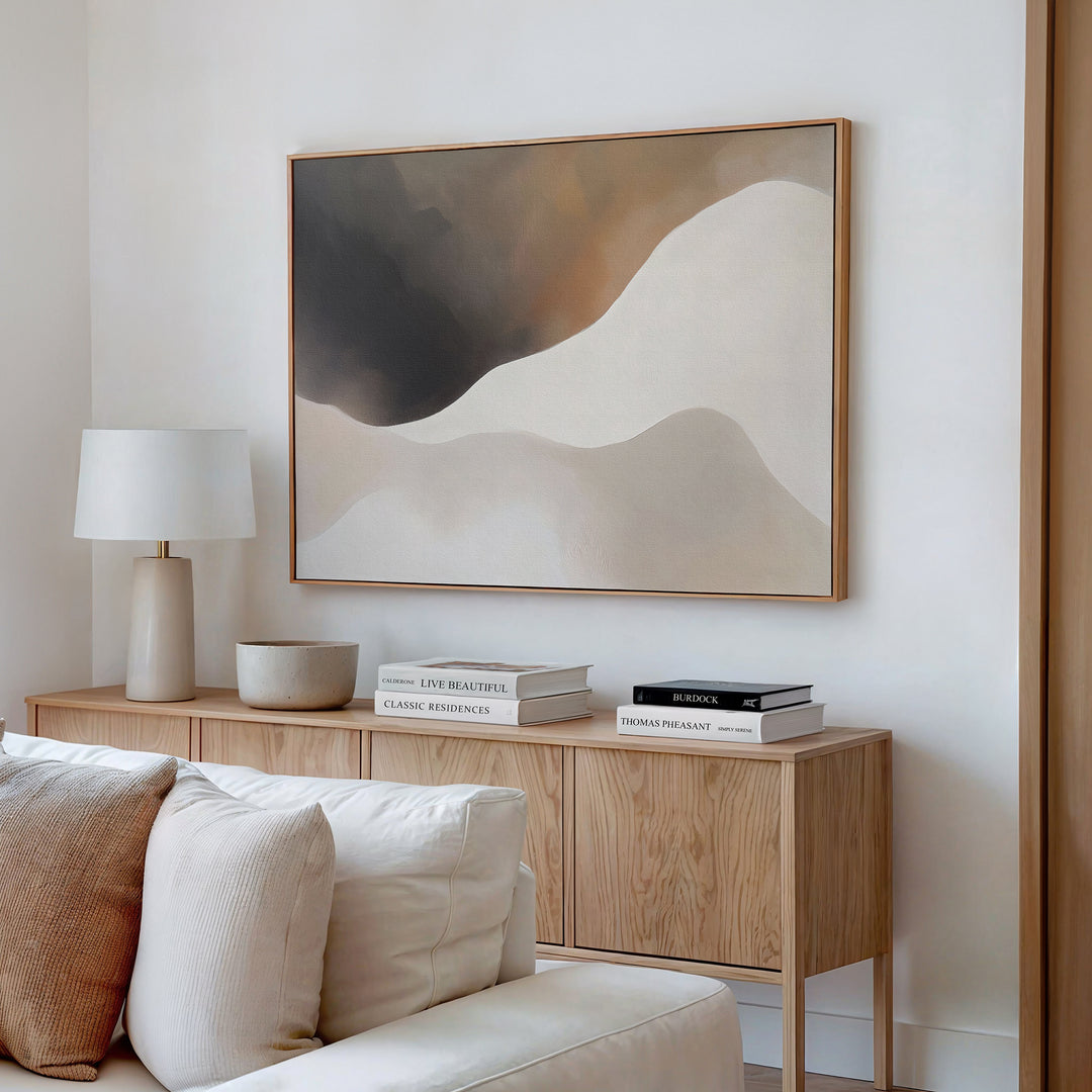 A modern living room featuring the Misty Silhouette Canvas Art – Horizontal, a light wood sideboard, white lamp, stacked books, bowl, and a beige sofa with cushions brings a refined touch to the neutral-toned decor.