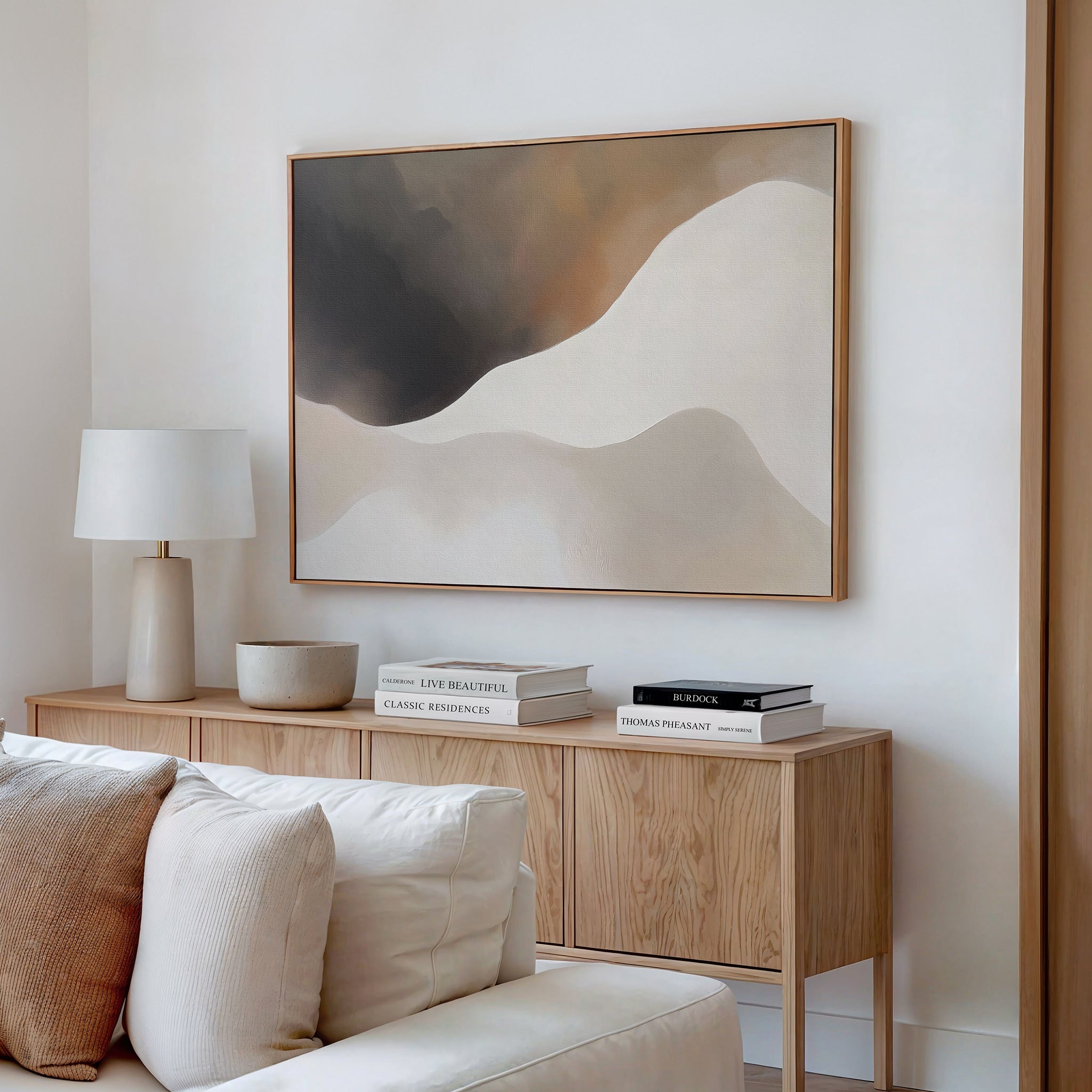 A modern living room featuring the Misty Silhouette Canvas Art – Horizontal, a light wood sideboard, white lamp, stacked books, bowl, and a beige sofa with cushions brings a refined touch to the neutral-toned decor.