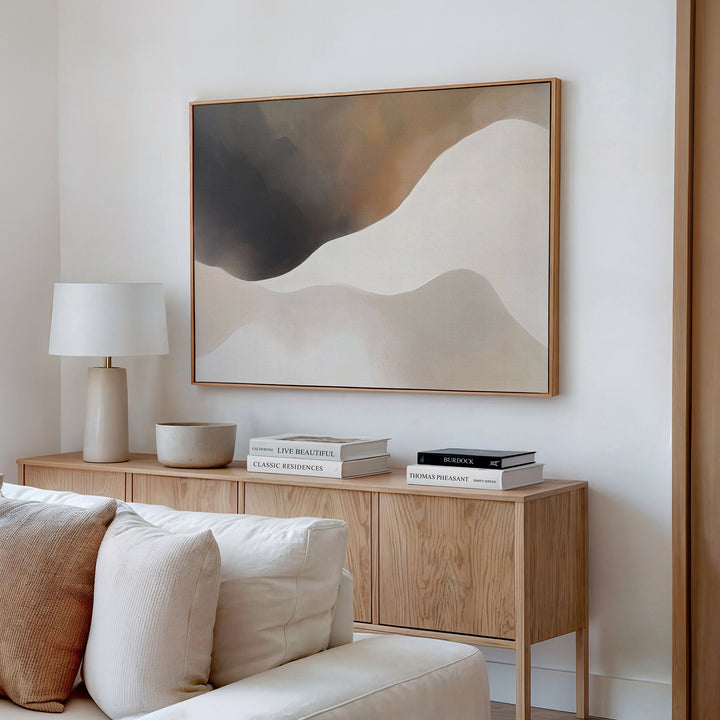 A modern living room featuring the Misty Silhouette Canvas Art – Horizontal, a light wood sideboard, white lamp, stacked books, bowl, and a beige sofa with cushions brings a refined touch to the neutral-toned decor.