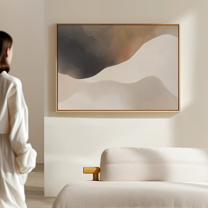 A woman in a white blouse stands in a minimal, neutral room, admiring the Misty Silhouette Canvas Art – Horizontal, featuring soft beige, brown, and cream wavy shapes above a light-colored sofa as modern living room decor.