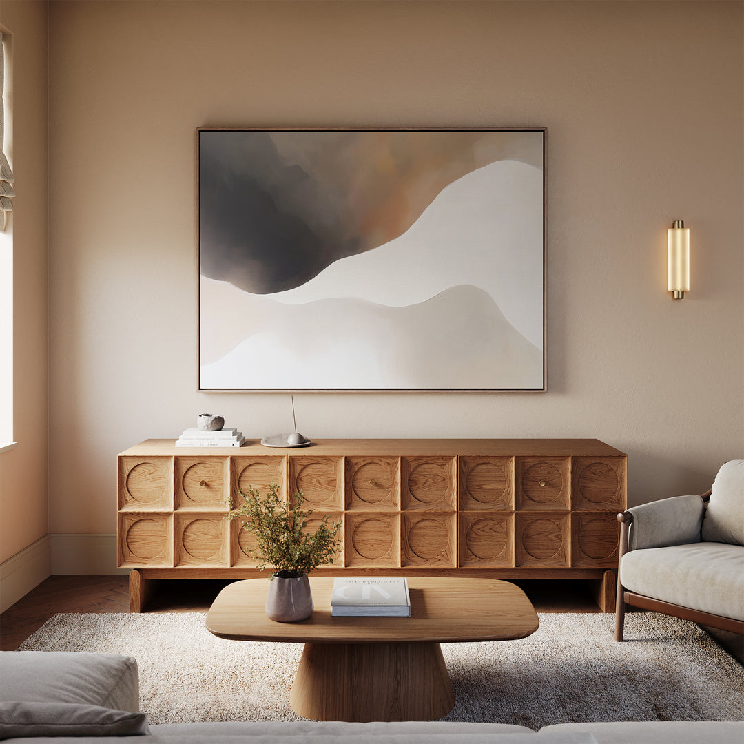 A modern living room with neutral tones features a wooden credenza with textured circles, the Misty Silhouette Canvas Art – Horizontal above it, a rounded coffee table with a book and vase, and a light sofa on a soft rug.