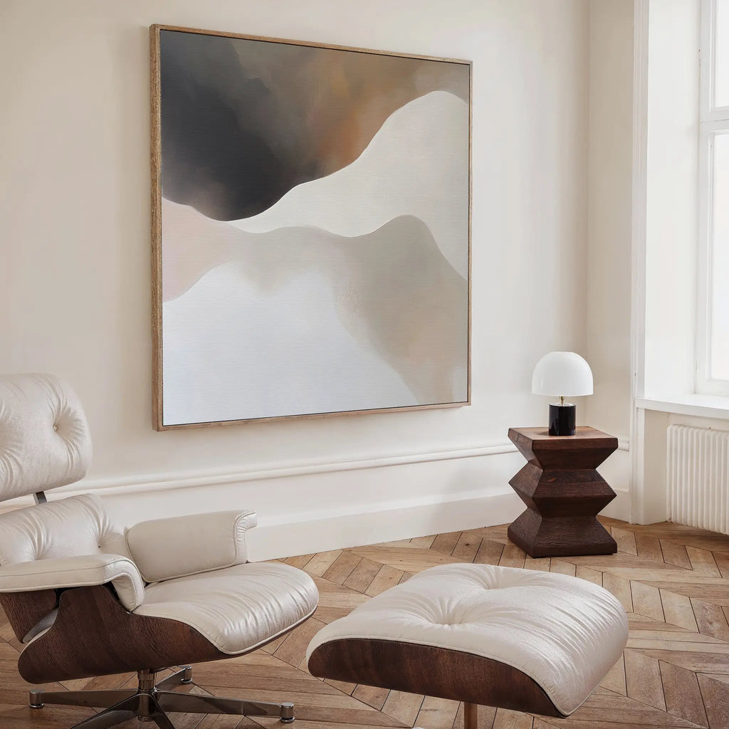 A modern living room with a cream leather lounge chair and ottoman on herringbone wood floors, a geometric dark wood side table with a white lamp, and Misty Silhouette Canvas Art in neutral tones for chic decor.