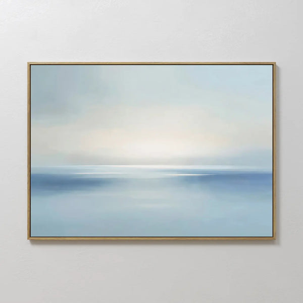 The Misty Waters Canvas Art features soft blue and white tones in a calm seascape design, accented by a thin gold frame for modern wall decor, perfect for displaying on a white wall.