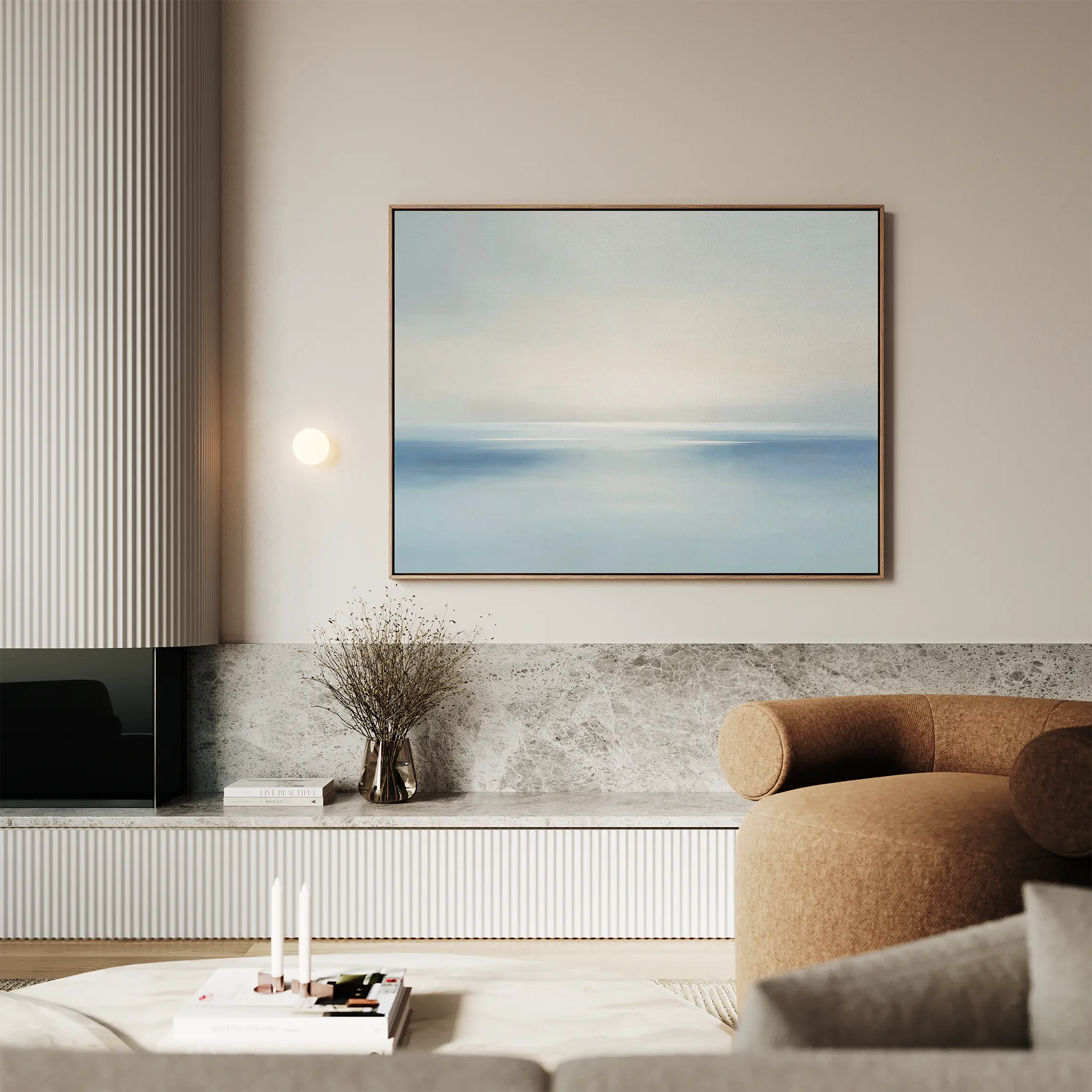 A modern living room features a curved beige sofa, marble accents, and serene decor. The Misty Waters Canvas Art—a soft abstract blue and white seascape—hangs above a built-in shelf with decor items and a small vase.