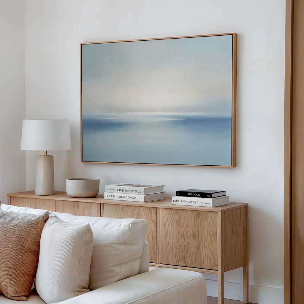 A minimalist living room features a light wooden sideboard, stacked books, a white lamp, a bowl, and serene Misty Waters Canvas Art—a large framed pastel seascape—hanging above a beige sofa on a white wall.