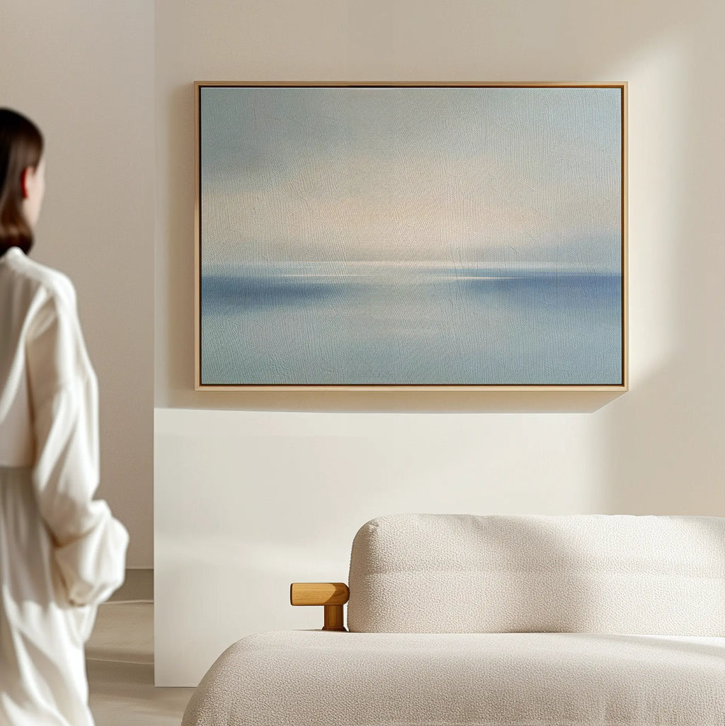 A person stands in a minimal, light-filled room, gazing at the Misty Waters Canvas Art with soft blue and beige tones hanging above a modern beige sofa.