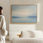 A person stands in a minimal, light-filled room, gazing at the Misty Waters Canvas Art with soft blue and beige tones hanging above a modern beige sofa.