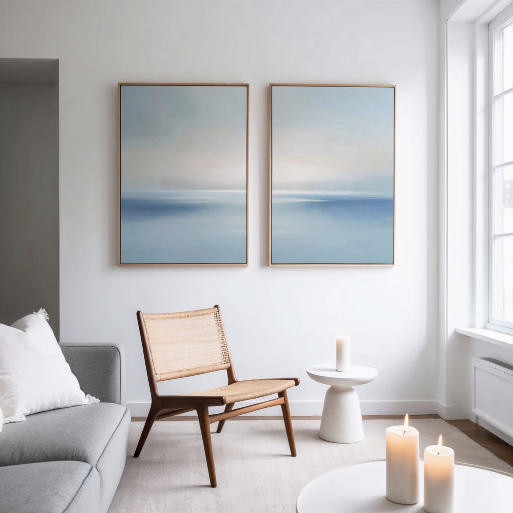 A modern, minimalist living room with a gray sofa, woven chair, small round side table, and three lit candles. The Misty Waters Set of 2 blue and white wall art hangs above the chair, with natural light streaming in.