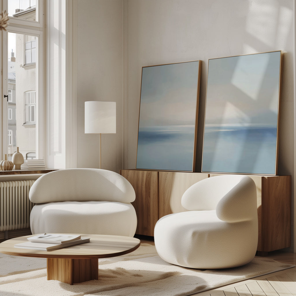 A minimalist living room with two modern white armchairs, a wooden coffee table, a floor lamp, and two Misty Waters Set of 2 abstract blue paintings leaning against a cabinet by a sunlit window.