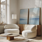 A minimalist living room with two modern white armchairs, a wooden coffee table, a floor lamp, and two Misty Waters Set of 2 abstract blue paintings leaning against a cabinet by a sunlit window.