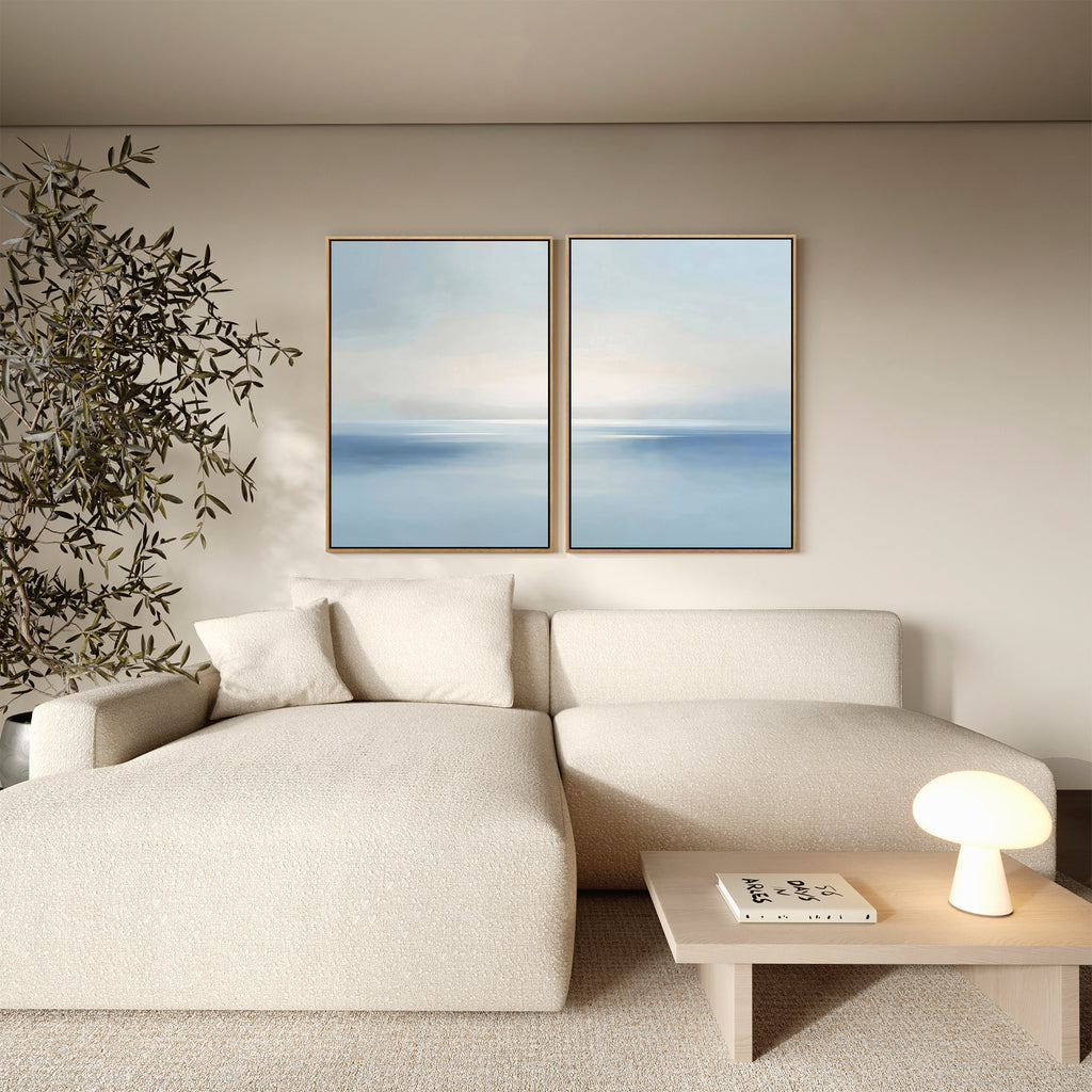 A modern living room with a cream sectional sofa, wooden coffee table with a book and lamp, large potted plant, and two Misty Waters Set of 2 serene wall art pieces on a beige wall.