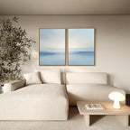 A modern living room with a cream sectional sofa, wooden coffee table with a book and lamp, large potted plant, and two Misty Waters Set of 2 serene wall art pieces on a beige wall.