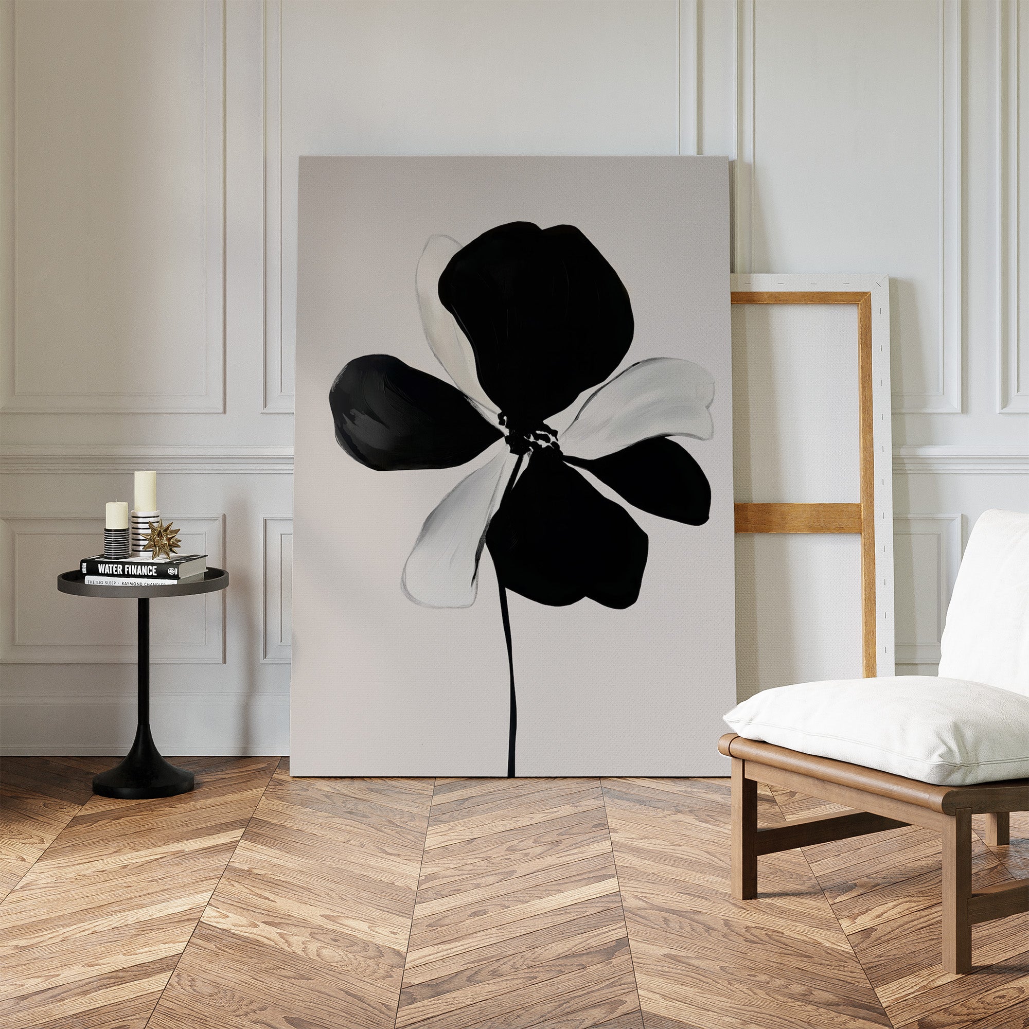 A minimal room with herringbone wood floors features the Monochrome Flora Abstract Canvas Art as minimalist wall decor, resting against a wall beside an empty frame, a white cushioned chair, and a small round table with candles and books.