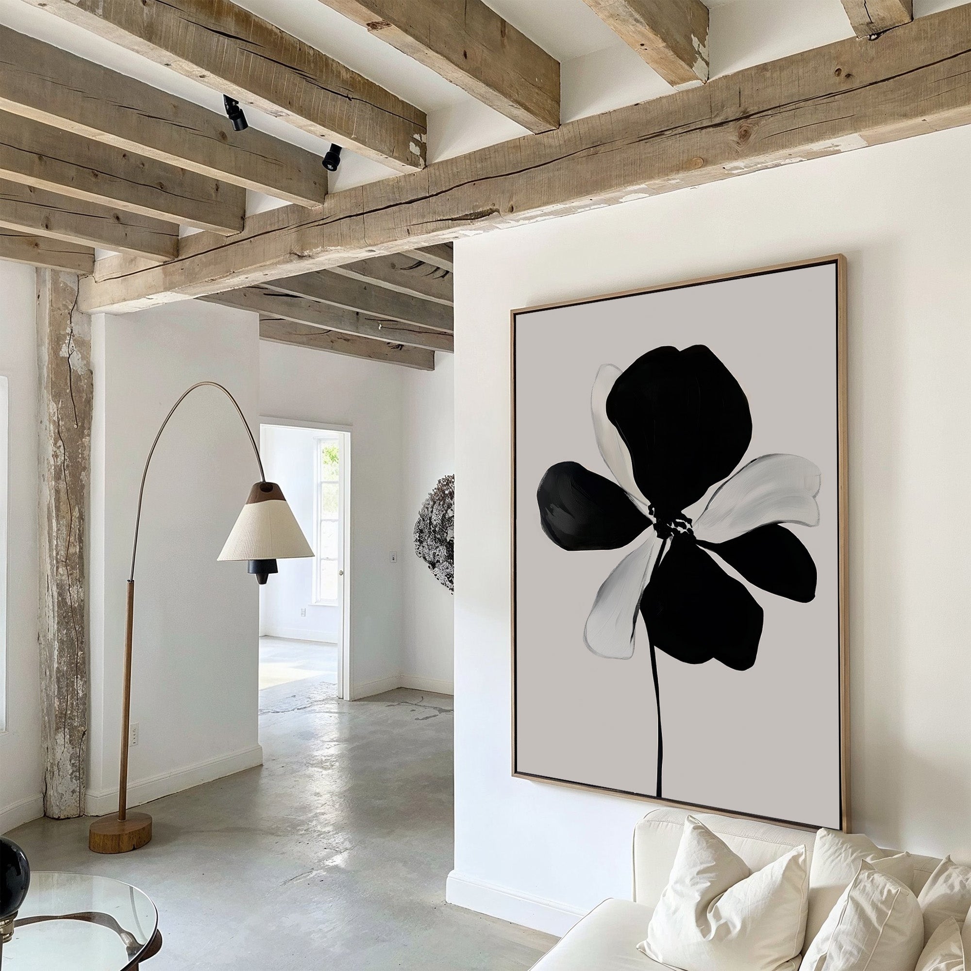 A modern living room features exposed wooden beams, white walls, a light sofa with cushions, a curved floor lamp, and the Monochrome Flora Abstract Canvas Art as minimalist wall decor. Natural light brightens the space.