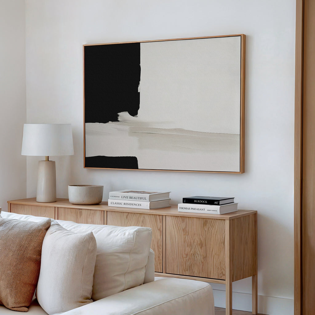 A minimalist living room corner featuring a wooden sideboard, books, a bowl, a white table lamp, and the Monochrome Harmony Canvas Art – Horizontal in black and beige above a white sofa.