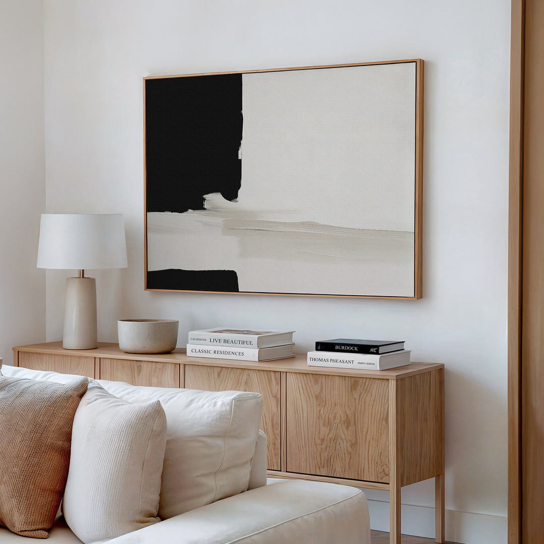 A minimalist living room corner featuring a wooden sideboard, books, a bowl, a white table lamp, and the Monochrome Harmony Canvas Art – Horizontal in black and beige above a white sofa.