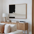 A minimalist living room corner featuring a wooden sideboard, books, a bowl, a white table lamp, and the Monochrome Harmony Canvas Art – Horizontal in black and beige above a white sofa.