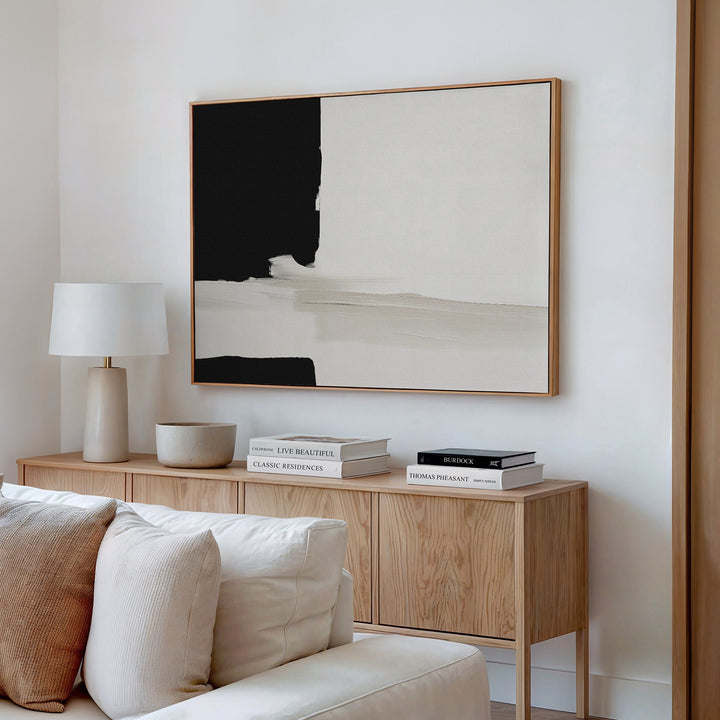 A minimalist living room corner featuring a wooden sideboard, books, a bowl, a white table lamp, and the Monochrome Harmony Canvas Art – Horizontal in black and beige above a white sofa.