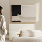 A woman stands in a minimal, modern room with a beige sofa. Soft natural light fills the space, highlighting the Monochrome Harmony Canvas Art – Horizontal framed on the wall.