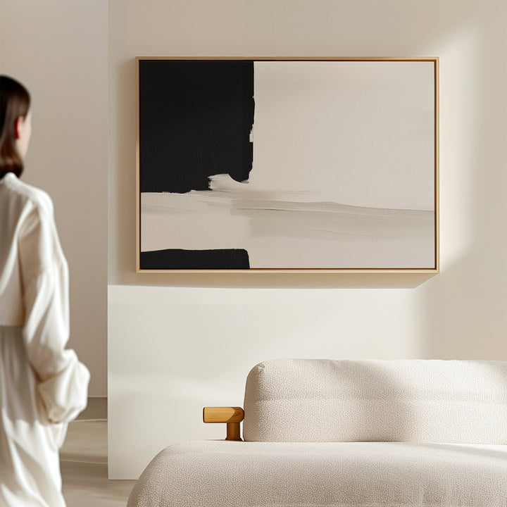A woman stands in a minimal, modern room with a beige sofa. Soft natural light fills the space, highlighting the Monochrome Harmony Canvas Art – Horizontal framed on the wall.