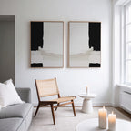 A minimalist living room features a light gray sofa, wooden chair, small white side table with a candle, and the Monochrome Harmony Set of 2 wall art. Sunlight streaming through tall windows enhances the space’s modern, clean lines.