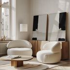 A modern living room features the Monochrome Harmony Set of 2 curved white armchairs, a round wooden coffee table, a wooden sideboard, and bold black and white wall art. Large windows fill the sunlit space with natural light.