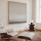 A modern, minimalist room features a cream Eames lounge chair with ottoman, a geometric wooden side table with dome-shaped lamp, herringbone wood floors, and the Morning Mist Abstract Canvas Art—a neutral-toned piece—on the wall.
