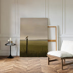 The Morning Perch Canvas Art, depicting a bird on a fencepost, leans against a white paneled wall and adds charm to this minimalist room with herringbone floors, earthy tones, a cushioned chair, and a side table with candles and books.