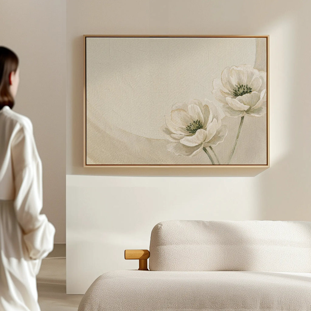A person in a white outfit stands in a minimalistic room, admiring the Morning Petals Canvas Art, which features two delicate white flowers and is displayed above a light-colored sofa on a beige wall.