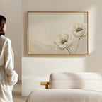 A person in a white outfit stands in a minimalistic room, admiring the Morning Petals Canvas Art, which features two delicate white flowers and is displayed above a light-colored sofa on a beige wall.