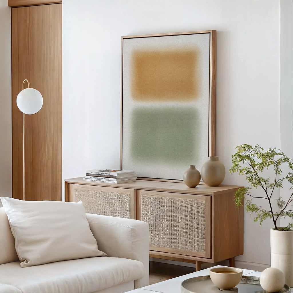 A minimalist living room with a light wooden sideboard, Morning Whisper Canvas Art featuring tan and green blocks, neutral vases, stacked books, a round glass coffee table, a soft white couch, and a leafy plant in the corner.