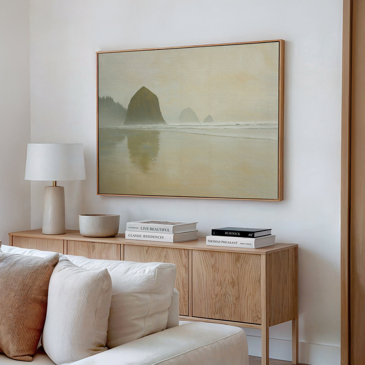 A minimalist living room features a wooden sideboard, stacked books, a lamp, and a round bowl. Above hangs the Morning At Cannon Beach Canvas Art for a coastal vibe. A cream sofa sits in the foreground.