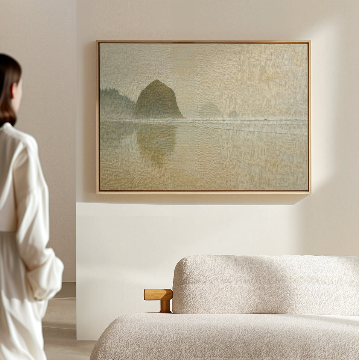 A person in white admires the Morning At Cannon Beach Canvas Art—a large, serene coastal print of rocks and calm water—displayed above a light-colored sofa in a minimal, softly lit room.