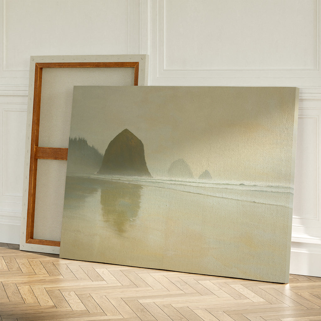 Two large canvases rest against a white wall on a wooden floor; one shows the back of a blank frame, while the other displays the Morning At Cannon Beach Canvas Art, featuring sea stacks reflected in wet sand.