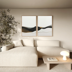 A modern living room with a cream sectional sofa, minimalist wooden coffee table, glowing table lamp, tall leafy plant, and the Mountain Contour Set of 2 abstract prints on the wall. The decor features neutral and earthy tones.