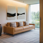 A modern living room features a tan leather sofa with three patterned pillows, Mountain Contour Set of 2 art above, a large window with sheer curtains, and a ceramic vase on checkered carpet. Trees are visible outside.