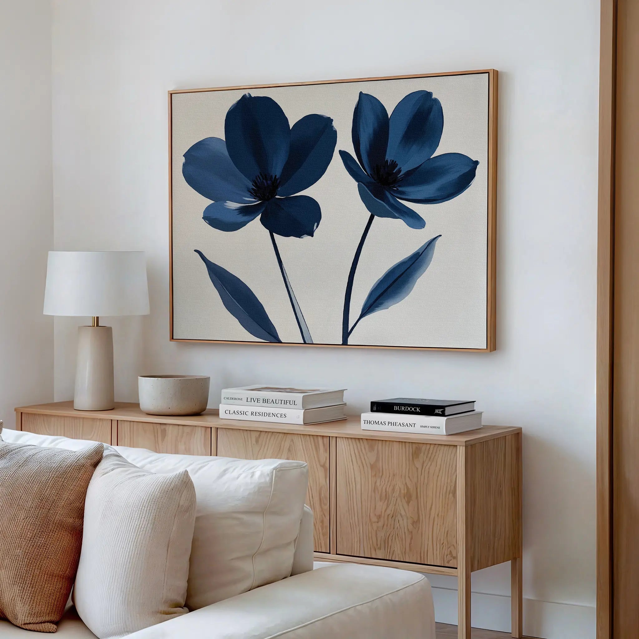 A modern living room features a light wood sideboard, white lamp, stacked books, round bowl, and Navy Flora Canvas Art—a minimalist wall decor with two blue flowers—above the sideboard. A cream sofa sits in the foreground.
