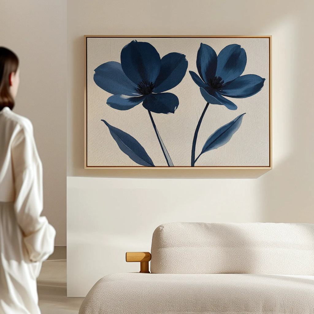 A person stands in a softly lit room, gazing at the Navy Flora Canvas Art—a framed minimalist print of two large blue flowers—displayed above a light-colored cushioned bench on a beige wall.
