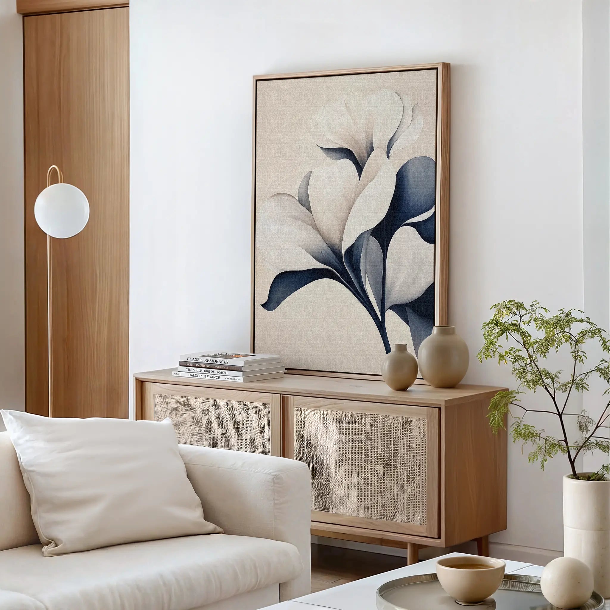 A modern living room with neutral decor, featuring a light wood sideboard, Navy Magnolia Canvas Art, ceramic vases, a potted plant, a white sofa with cushions, and a round pendant light.