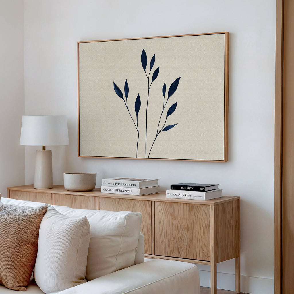 A minimalistic living room with a light wood sideboard, stacked books, a white lamp, and the Navy Stem Canvas Art featuring black botanical shapes above a white sofa with cushions.