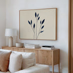 A minimalistic living room with a light wood sideboard, stacked books, a white lamp, and the Navy Stem Canvas Art featuring black botanical shapes above a white sofa with cushions.