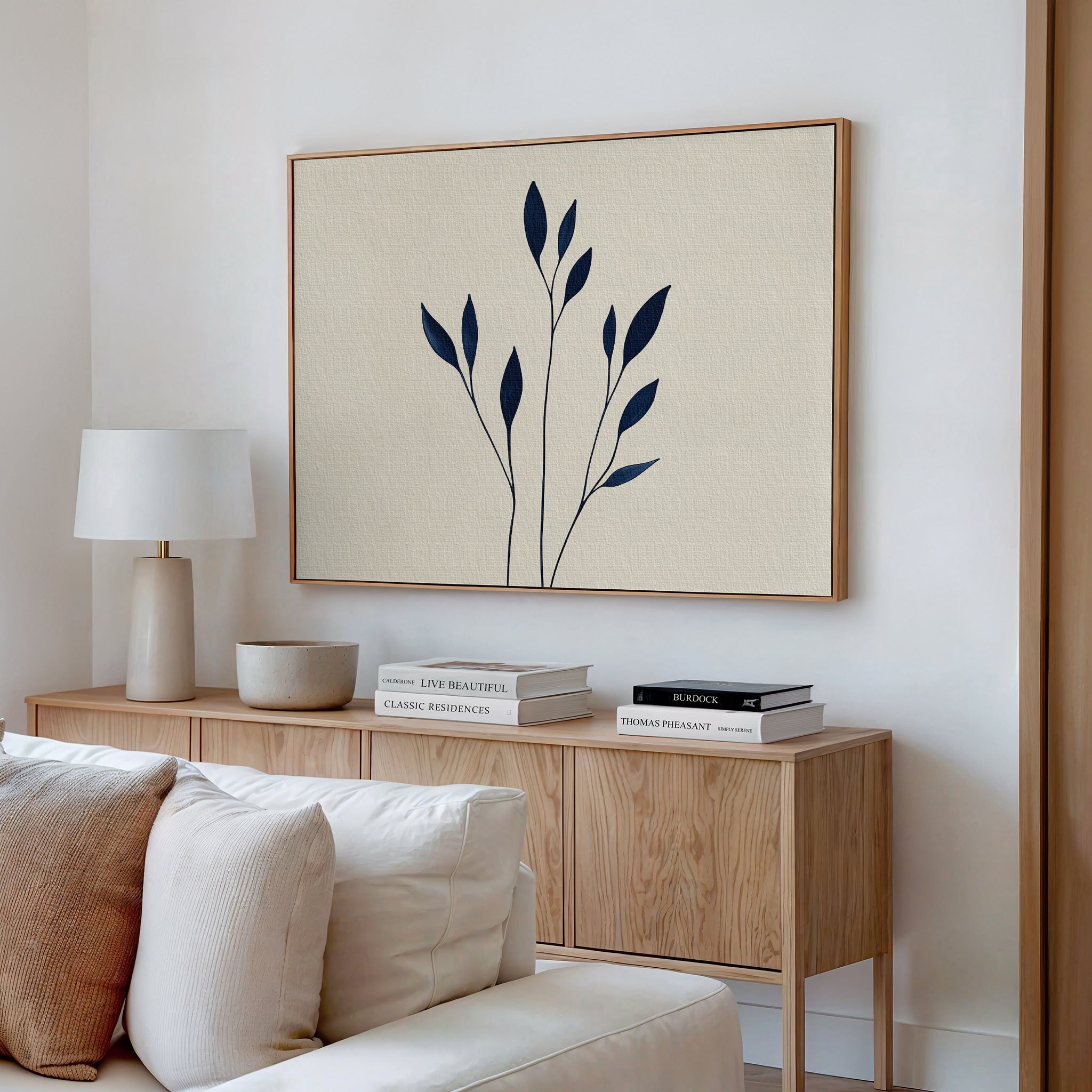 A minimalistic living room with a light wood sideboard, stacked books, a white lamp, and the Navy Stem Canvas Art featuring black botanical shapes above a white sofa with cushions.