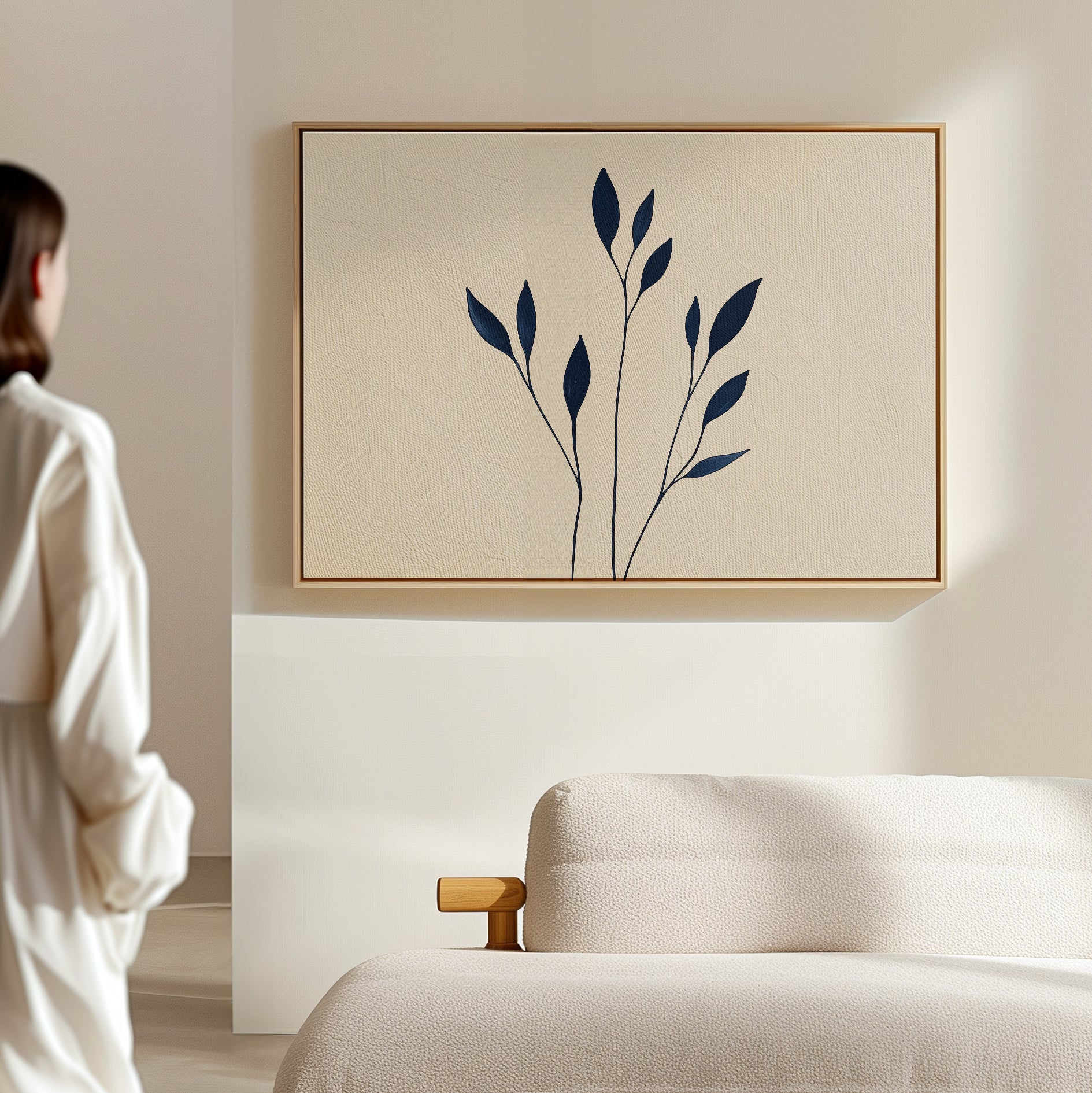 A person stands in a bright, minimal room facing a beige wall, where the Navy Stem Canvas Art with bold dark blue shapes is displayed above a cream-colored sofa.