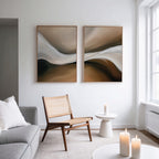 A modern living room with neutral decor, including a wicker chair, light gray sofa, small white table with candles, and the Neutral Landscape River Set of 2 minimalist wall art in brown and white tones. Natural light streams through large windows.