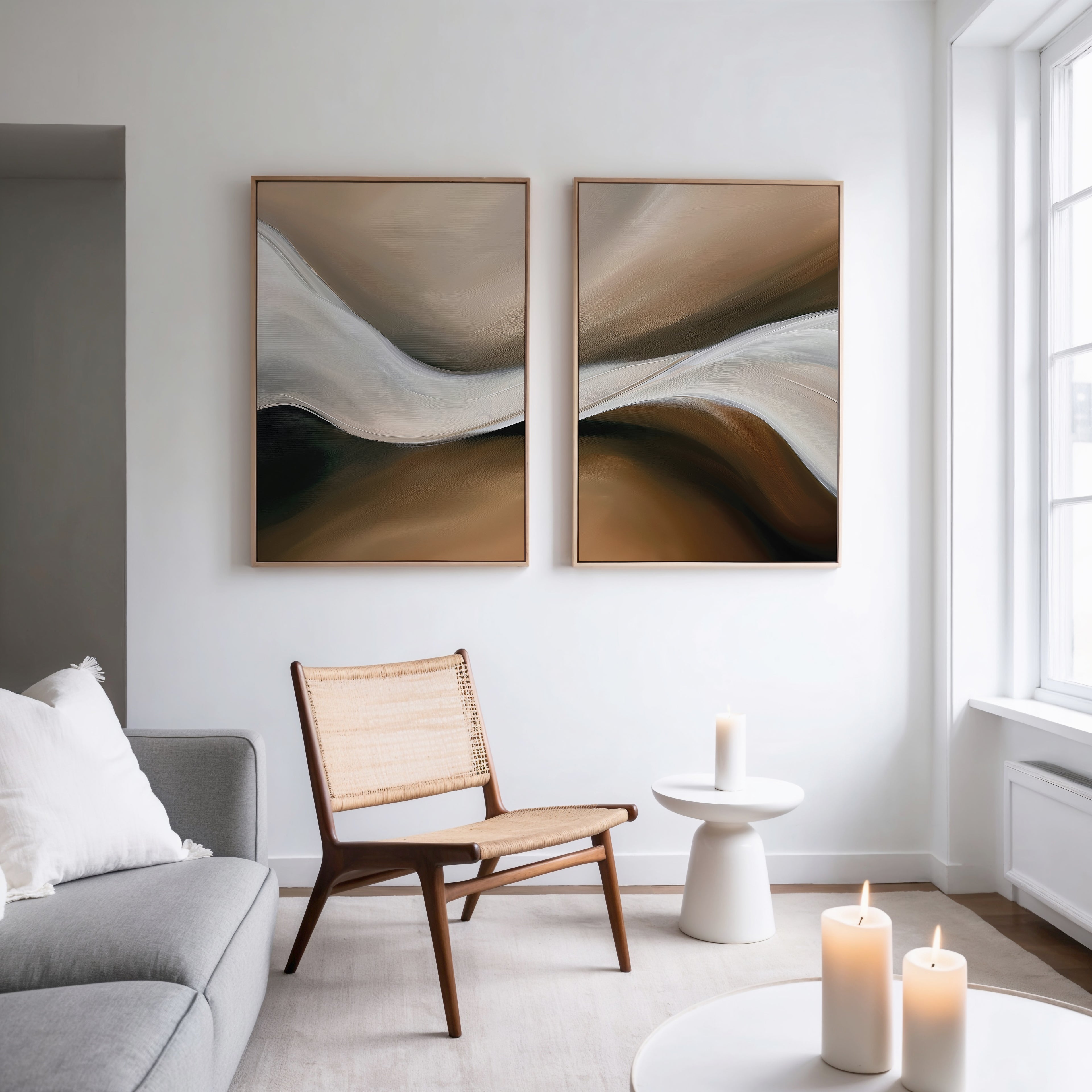 A modern living room with neutral decor, including a wicker chair, light gray sofa, small white table with candles, and the Neutral Landscape River Set of 2 minimalist wall art in brown and white tones. Natural light streams through large windows.