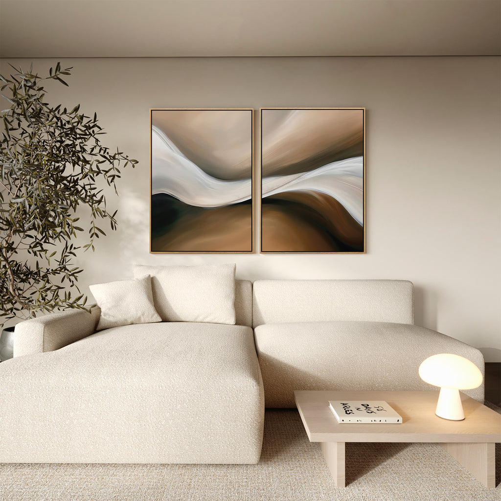 A modern living room with a cream-colored sectional sofa, featuring the “Neutral Landscape River Set of 2” minimalist wall art above, a small light wood table with a round lamp and book, and a tall leafy plant in the corner.