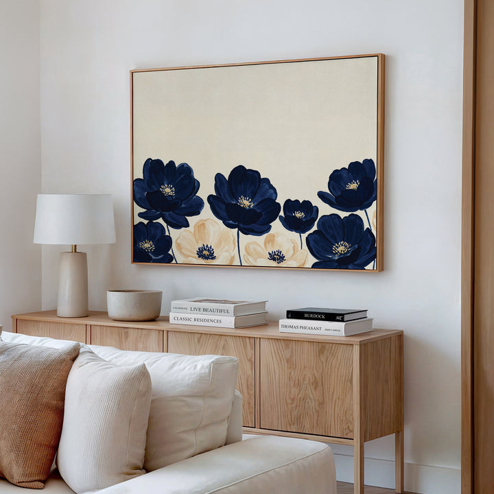 A modern living room features a light wood sideboard with a lamp, decor, and books. Above it hangs the Nightshade Canvas Art – Horizontal in navy and beige. A cream sofa appears in the foreground.