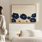 A person stands in a minimalist room, admiring the Nightshade Canvas Art – Horizontal, featuring large blue and beige flowers framed on a light-colored wall above a cream sofa.