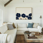 A cozy living room features a white sectional sofa, assorted pillows, a wooden coffee table, and the Nightshade Canvas Art – Horizontal with blue and white floral accents. White shiplap walls and rustic decor complete the inviting space.