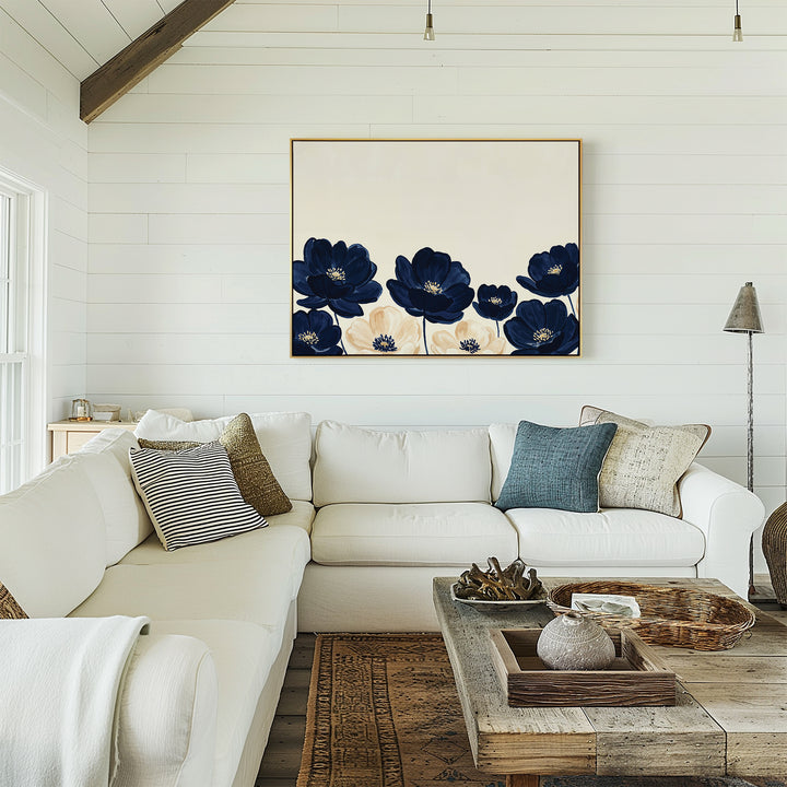A cozy living room features a white sectional sofa, assorted pillows, a wooden coffee table, and the Nightshade Canvas Art – Horizontal with blue and white floral accents. White shiplap walls and rustic decor complete the inviting space.